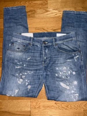 Blå slitna Dondup jeans med raka ben - Säljer ett par blå jeans med slitningar och raka ben. Jeansen har klassisk femficksmodell, knappgylf och ljus tvätt med tydliga slitdetaljer framtill och baktill. Perfekta för en avslappnad och trendig look. Knappast använda. 