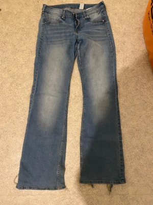 Blå bootcut jeans med slitna detaljer - Ett par klassiska blå bootcut jeans med slitningar vid bensluten och lätt tvättade partier framtill och baktill. Håll vid fickan men snus knappt. Perfekta för en avslappnad och trendig look.
