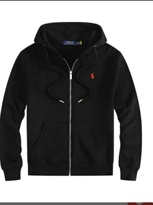 Svart hoodie från Polo Ralph Lauren - Snygg svart hoodie med dragkedja från Polo Ralph Lauren. Klassisk design med huva, fickor framtill och det ikoniska röda Polo-logot på bröstet. Tillverkad i mjukt bomullsmaterial och har ribbade muddar vid ärmar och nederkant.