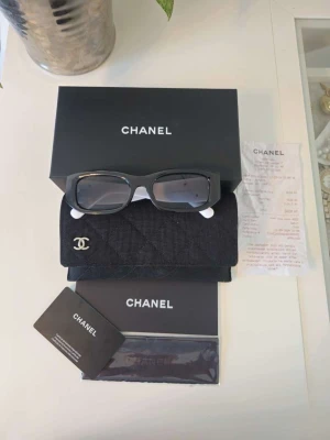 Chanel svarta solglasögon med vita skalmar - Snygga solglasögon från Chanel med svart rektangulär front och vita skalmar med silverfärgad CC-logga. Kommer med originalfodral, box och äkthetskort. Perfekt accessoar för dig som vill sticka ut med lyxig stil.