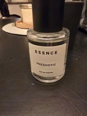 ESSNCE Freshotic Eau de Parfum - Minimalistisk parfymflaska från ESSNCE med doften Freshotic. Flaskan är genomskinlig med en vit etikett och svart text, samt ett stort svart lock. Modern och stilren design som passar perfekt på parfymhyllan.