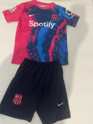FC Barcelona Nike matchställ barn - Säljer ett FC Barcelona matchställ för barn från Nike. Tröjan är rosa och blå med klubbmärke, sponsorlogga och tryck, medan shortsen är mörkblå med klubbmärke och Nike-logga. Perfekt för fotbollsträning eller att heja på laget i.