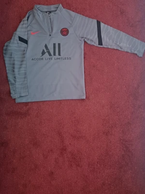 Grå PSG träningströja Nike - Grå långärmad träningströja från PSG med svart och röd logga, halv dragkedja och svarta detaljer på ärmarna. Materialet är troligtvis polyester, perfekt för sport och träning. Tryck med 'Accor Live Limitless' och 'Visit Rwanda' på ryggen.