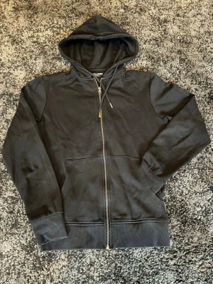 Svart zip-hoodie med huva från Jack&Jones  - Svart hoodie med dragkedja från Jack&Jones. Klassisk modell med huva och dragsnören, samt två stora fickor framtill. Ribbstickade muddar vid ärmslut och nederkant. Perfekt för en avslappnad och stilren look.