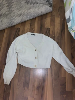 Vit cropped kofta med knappar - Snygg vit cropped kofta med stora knappar framtill och v-ringad hals. Koftan har långa ärmar och är stickad i ett mjukt material. Perfekt att slänga över en topp för en chill look.