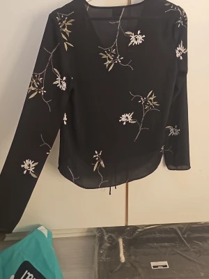 Svart blus med blommigt mönster - Snygg svart blus med vita och beige blommor, v-ringning och dragsko framtill. Lätt transparent material som ger en luftig känsla. Långa ärmar och avslappnad passform, perfekt till jeans eller kjol. Märket syns inte tydligt på bilderna.