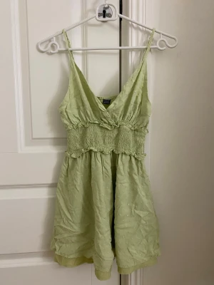 Ljusgrön kort klänning med volang - Söt ljusgrön kort klänning med smala axelband och v-ringning. Klänningen har smock i midjan, volangdetaljer och ett lätt, luftigt tyg som ger en somrig känsla. Perfekt för varma dagar och enkel att styla med accessoarer.