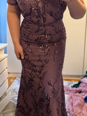 Lång lila paljettklänning med spets - Säljer en elegant långklänning i lila med glittriga paljetter och spetsdetaljer. Klänningen har korta ärmar, v-ringning och ett vackert blommigt mönster. Perfekt för bal eller festliga tillfällen.