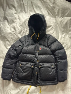 Svart  Fjällräven Expedition - Svart dunjacka från Fjällräven  Expedition Series med stor huva, gula dragskor och två stora framfickor med tryckknappar. Stl M passar 170-175 