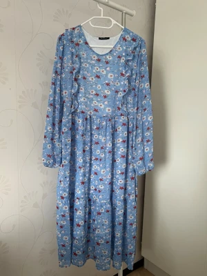 Blommig blå långklänning - Söt blå långklänning med blommigt mönster i vitt och rött. Klänningen har långa ärmar, rund halsringning och volangdetaljer framtill. Tillverkad i ett lätt och luftigt material, perfekt för varmare dagar. Är köpt utomlands 💙har L i storlek men passar också M då de är en band man kan dra i midjan