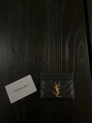 Svart YSL kortfodral i läder - Stilrent svart kortfodral från Yves Saint Laurent i präglat läder med ikoniskt guldigt YSL-emblem framtill. Fodralet har flera kortfack och en smidig, tunn design som passar perfekt i fickan. Lyxig accessoar för dig som gillar designerplånböcker.