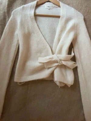 Krämvit omlottkoft  - krämvit omlottkofta med långa ärmar och knytband i midjan. Koftan har en mjuk, stickad känsla och en enkel, stilren design som passar perfekt till jeans eller kjol.