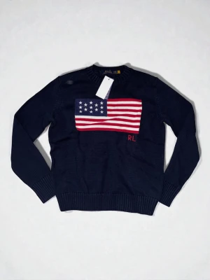 Mörkblå stickad tröja Polo Ralph Lauren - Snygg mörkblå stickad tröja från Polo Ralph Lauren med amerikansk flagga i rött, vitt och blått på bröstet samt RL-broderi. Tröjan har rund halsringning och ribbade muddar vid ärmslut och nederkant. Perfekt för dig som gillar klassisk preppy stil.