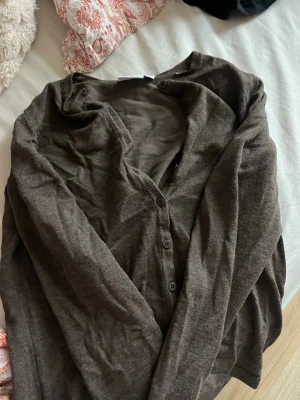 Mörkbrun tunn kofta med knappar - Tunn och mjuk mörkbrun kofta med långa ärmar och tre knappar framtill. Perfekt att slänga över en t-shirt eller linne. Enkel och stilren design utan synliga loggor eller mönster.