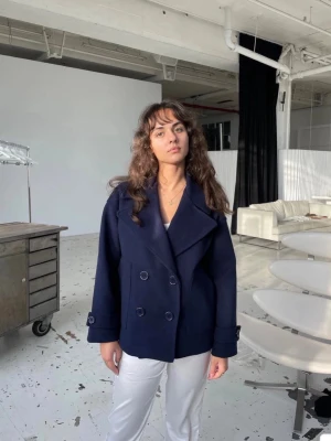 Mörkblå dubbelknäppt kappa - Snygg mörkblå kappa med dubbelknäppning och bred krage. Jackan har långa ärmar och knappar på ärmsluten. Passar perfekt för dig som vill ha en stilren och klassisk look. Materialet ser ut att vara ullblandning. Passar M/L