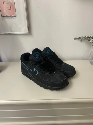 Nike Air Max 90 svart/blå sneakers - Svarta Nike Air Max 90 sneakers med blå detaljer och logga. Klassisk siluett med mesh och syntetmaterial, robust sula och ikonisk Air Max-dämpning. Snygg kontrast mellan svart och blått gör dem riktigt fräscha till streetwear.