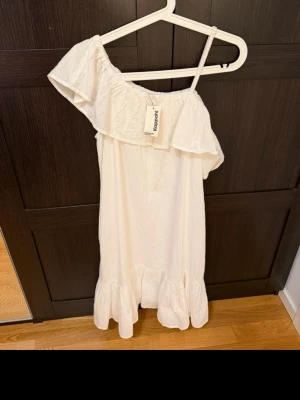 Klänning - Söt vit klänning med volangdetaljer och en axelbandslös design på ena sidan. Klänningen har en luftig passform och är perfekt för varma dagar. Tillverkad i ett lätt bomullsmaterial och har en kort längd med volang nertill.