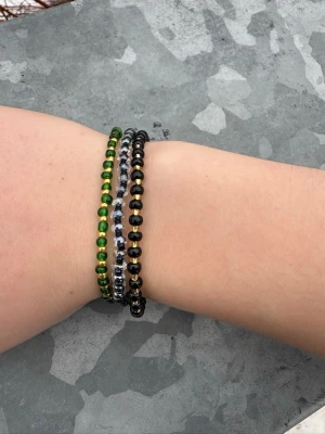 Set med tre färgglada pärlarmband - Säljer ett set med tre elastiska armband gjorda av små pärlor. Ett armband är svart och guld, ett är grönt och guld, och det tredje är svart och vitt. Perfekta för att mixa och matcha till olika outfits. Alla armband är handgjorda och har en enkel, stilren design.