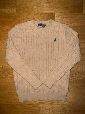 Beige kabelstickad tröja Polo Ralph Lauren - Kabelstickad beige tröja från Polo Ralph Lauren med rund halsringning och mörkblå broderad logga på bröstet. Tröjan har ribbade muddar vid ärmslut och nederkant. Perfekt för lager-på-lager och klassisk stil. Hör av er vid intresse. Priset går att diskutera.