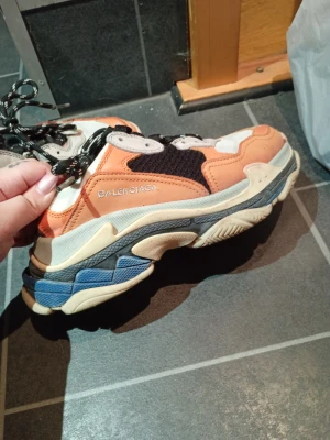 Balenciaga Triple S sneakers orange - Chunky Balenciaga Triple S sneakers i orange, svart, vitt och blått. Skorna har en flerskiktad sula, mesh- och läderdetaljer samt broderad logga på sidan. Svarta och vita snören ger en sportig vibe.