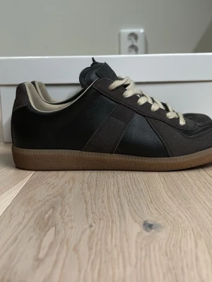 Svarta Maison Margiela Replica sneakers - Maison Margiela Replica sneakers i svart läder och mocka med beige detaljer och gummisula. Klassisk låg modell med beige snörning och vit innersula. Snygg retrostil som passar till det mesta och ger en clean look. Endast använda ett fåtal gånger.