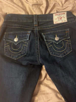 Mörkblå True Religion jeans - Säljer ett par mörkblå jeans från True Religion med ikoniska kontrastsömmar och snygga fickdetaljer med lock och silverknappar. Klassisk femficksmodell i denim med gul och orange söm. Perfekta för dig som gillar streetwear och vill sticka ut.