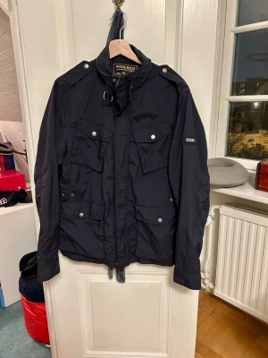 Woolrich fieldjacket mörkblå - Snygg svart fieldjacket från Woolrich med fyra fickor där fram. Jackan har hög krage med spänne och diskret logga på ärmen. Perfekt nu till våren och kallare sommarkvällar.