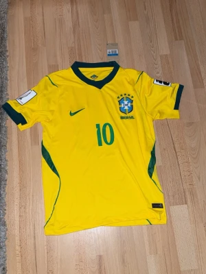 Brasilien Neymar Jr matchtröja gul - Säljer en gul Brasilien fotbollströja från Nike med Neymar Jr och nummer 10 på ryggen. Tröjan har gröna detaljer, officiellt CBF-märke och Nike-logga på bröstet. Perfekt för dig som älskar fotboll och vill stötta Brasilien på matchdagar.