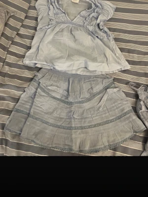 Ljusblå volangblus med spetsdetaljer - Supersöt ljusblå blus med volanger och spetsdetaljer. V-ringad framtill och ärmlös design som ger en luftig känsla. Perfekt för varma dagar och enkel att matcha med kjol eller jeans. 250 för vardera plagg 