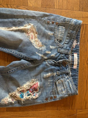 Blå slitna jeans med patchdetaljer - Jättecoola Vero Moda jeans med slitningar och färgglada patchdetaljer under de trasiga partierna. Midjemåttet är 38 cm och innerbenslängden är 77 cm.