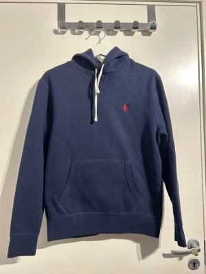 Mörkblå hoodie från Ralph Lauren - Mörkblå hoodie från Ralph Lauren med vit dragsko i huvan och klassisk känguruficka framtill. Liten röd Polo-logga broderad på bröstet. Tillverkad i mjukt bomullsmaterial och har ribbade muddar vid ärmslut och nederkant.