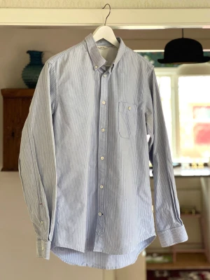 Blåvit randig skjorta från NN07 - Klassisk blåvit randig skjorta från NN07 i bomull. Modell Derek. Skjortan har lång ärm, bröstficka med knapp och vita knappar framtill. Perfekt till jeans eller chinos för en clean och stilren look. Endast använd ett par gånger så skjortan är i nyskick. 
