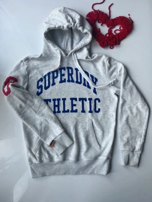 Grå Superdry hoodie med tryck - Snygg ljusgrå hoodie från Superdry med blått 'SUPERDRY ATHLETIC'-tryck på bröstet och rött 98-tryck på ena ärmen. Klassisk huva med dragsko och känguruficka framtill. Perfekt för en sportig och avslappnad stil.