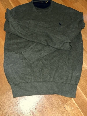 Mörkgrön tröja från Polo Ralph Lauren - Mörkgrön stickad tröja från Polo Ralph Lauren med rund halsringning och diskret blå logga broderad på bröstet. Tröjan har långa ärmar och ribbade muddar vid ärmslut och nederkant. Perfekt för lager på lager och enkel att matcha.