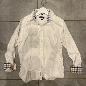 Vit linneskjorta - Klassisk vit skjorta från Tom Rusborg of Copenhagen i linne med lång ärm och rutiga detaljer i blått och beige på insidan av manschetterna och kragen. Skjortan har knappar framtill och en avslappnad passform. Perfekt för en stilren look.