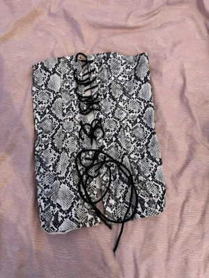 Korsett med ormmönster och snörning - Säljer en snygg korsett med svartvitt ormmönster och snörning framtill. Korsetten har hakar för stängning baktill och är tillverkad i ett glansigt tyg. Perfekt för att ge outfiten en edgy touch.