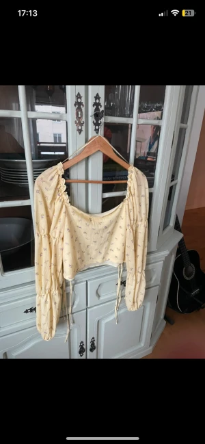 Gul blommig blus  - Söt gul blus med små lila blommor och puffiga långärmar. Blusen har snörning baktill samt rynkade detaljer vid axlar och ärmslut. Tillverkad i ett lätt tyg som känns luftigt och somrigt. 