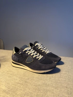 Philippe Model blå sneakers - Snygga sneakers från Philippe Model i marinblå mocka och textil med vita snören och silverdetalj på hälen. Klassisk låg modell med rund tå och vit sula. Perfekta till jeans eller shorts för en clean och sportig look.