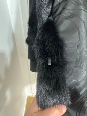 Svart päls kappa - Snygg svart kappa i päls med rak passform och mjukt foder. Kappan har en klassisk krage och stängs med diskreta hakar framtill. Perfekt för dig som vill ha en lyxig look och hålla dig varm under kalla dagar.