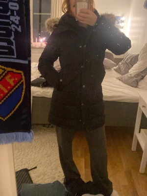 Svart lång dunjacka med päls - Snygg svart lång dunjacka med huva och päls. Jackan har knappar och dragkedja framtill samt två fickor på sidorna. Perfekt för kalla vinterdagar och ger en stilren look. Passar dig som vill ha både värme och stil.