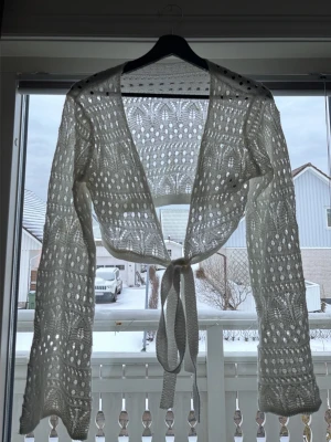 Vit virkad knytkofta - Säljer en vit virkad kofta med knytband framtill. Koftan har ett öppet hålmönster, vida ärmar och är croppad i modellen. Perfekt för lager på lager och ger en bohemisk vibe till din outfit.