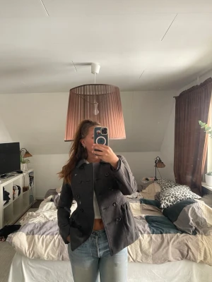 Mörkgrå kort kappa med knappar - Snygg mörkgrå kappa med dubbelknäppning och klassisk krage. Kappan har två fickor framtill och är tillverkad i ett mjukt tyg som känns bekvämt.