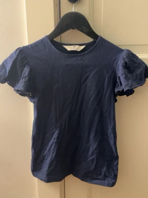 Mörkblå topp med volangärmar från H&M - Söt mörkblå topp från H&M med korta volangärmar i broderad bomull. Perfekt till jeans eller kjol för en trendig look. Klassisk rund halsringning och mjukt material som känns skönt mot huden. Storlek 170 men sitter som en vanlig xs🥰