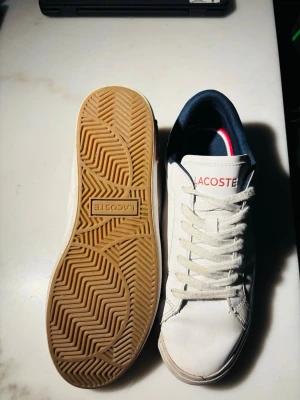 Vita Lacoste sneakers  - Snygga vita sneakers från Lacoste med klassisk krokodil-logga på sidan och röda samt blå detaljer. Skorna har vita snören, mörkblå insida och en slitstark gummisula med mönster för bra grepp. Perfekta för en clean och sportig look. De är i storlek 42 men passar 43!