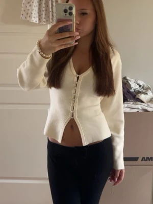 Vit kofta med pärlknappar - Trendig vit kofta med djup v-ringning och snygga pärlknappar framtill. Modellen är figurnära med lång ärm och lätt utsvängd nederkant. Perfekt att styla med jeans eller kjol för en chic look.