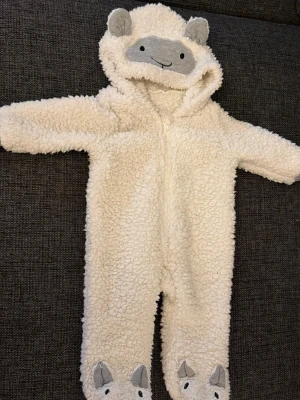 Vit teddyfodrad overall med huva - Supermjuk och fluffig vit overall i teddy-material med dragkedja framtill. Huvan har söta öron och ett broderat ansikte, och fötterna har gulliga detaljer som liknar djuransikten. Perfekt för kalla dagar när du vill vara extra varm och mysig.