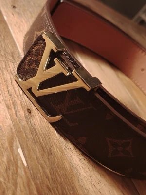 Louis Vuitto  - Säljer ett klassiskt brunt Louis Vuitto skärp med ikoniskt monogrammönster och stor L*-logga i guld som spänne. Bältet är gjort i skinn och har snygga detaljer som gör det till en riktig statement-accessoar.