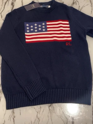 Ralph lauren usa tröja  - Mörkblå stickad tröja från Ralph Lauren med amerikansk flagga i rött, vitt och blått på bröstet. Tröjan har rund halsringning, ribbade muddar och RL-broderi under flaggan. Perfekt för dig som gillar klassisk preppy stil.