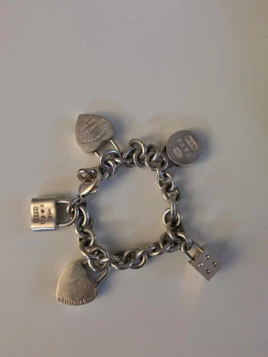 Tiffany & Co vintage  - Äkta silver armband från Tiffany & Co med chunky länkar och fem olika berlocker: två hjärtan, ett lås, en tärning och en rund bricka. Berlockerna har sina äkta Tiffany & Co stämplar. En Tiffany Sterling Silver Circle Link Chain från den berömda "Return to Tiffany"-kollektionen med 5 berlocker (tre av de är vintage och mycket sällsynta). En fantastisk klassisk vintage Tiffany & Co kedjearmband i sterling silver, välanvänd men fortfarande en klassiker för den som uppskattar det här fina märket. 
