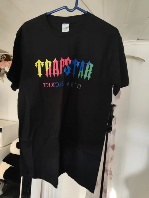 Trapstar svarta shorts med regnbågstryck med t-shirt sett - Svarta shorts med t-shirt från Trapstar med färgglatt regnbågstryck och texten 'IT'S A SECRET' på vänster ben. Shorts med elastisk midja och snörning för skön passform. Perfekta för chill dagar och streetwear-stil. Ta en storlek större 